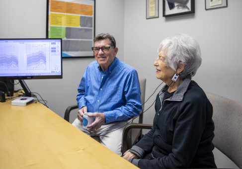An older couple in an audiology clinic reviews test results on a monitor. The woman is wearing a wired in-ear testing device